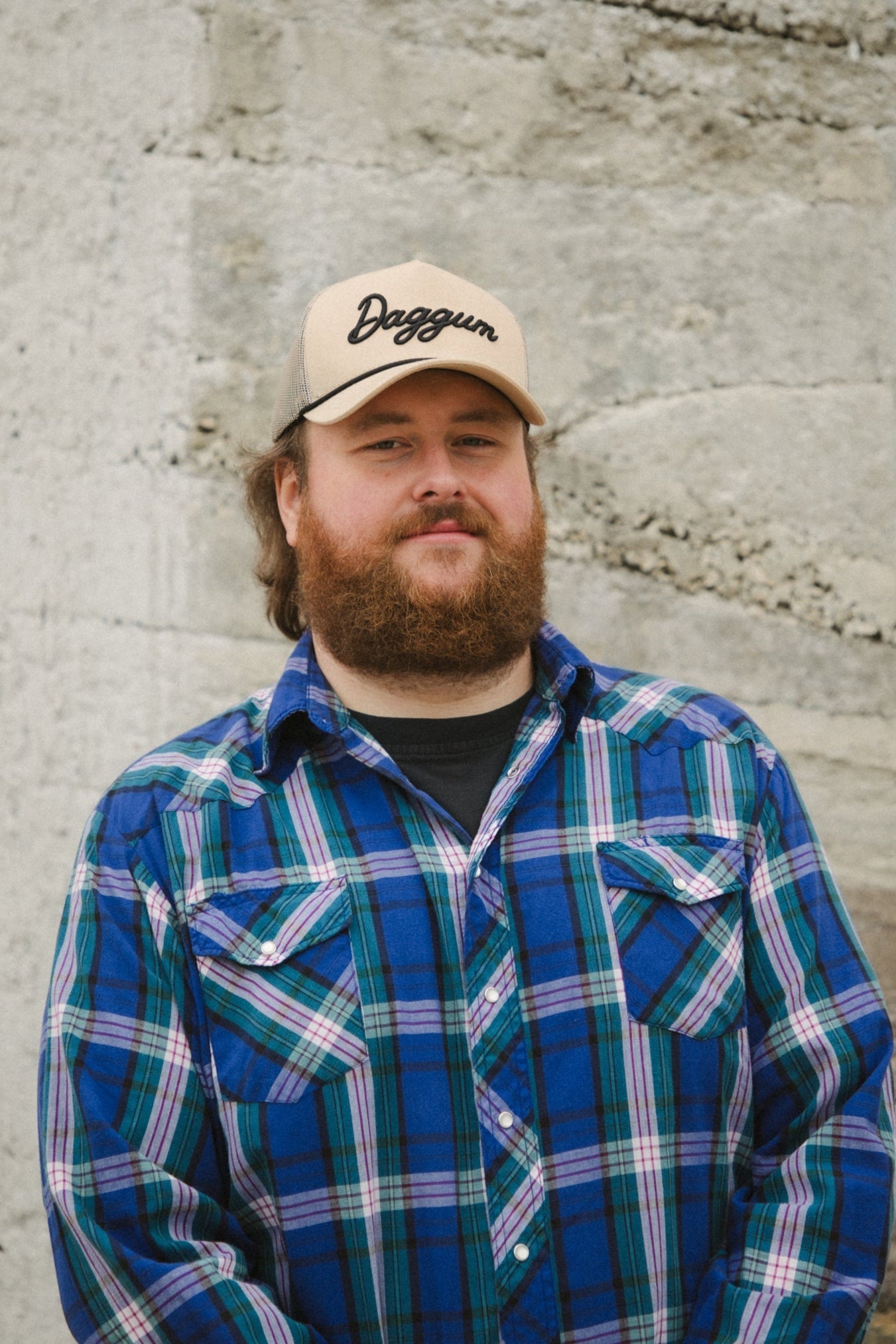 Man wearing a plaid shirt and cap with 'Dagwood' logo against a textured wall.
